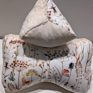 Op de afbeelding: Tweedelige kussenset met een botanische print van wilde bloemen en vlinders in bruin, oranje en geel op een witte achtergrond. Het bovenste kussen is driehoekig met een pluizige crèmekleurige rand. Het onderste kussen is een gebogen bolster.