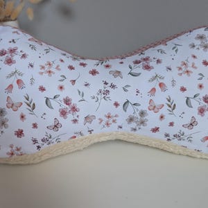Op de afbeelding: Een botvormig kussen met een wit bloemenpatroon met roze bloemen en vlinders. Het kussen heeft een zachte, crèmekleurige rand en een roze bies. Het kussen is ontworpen voor comfort en ondersteuning.