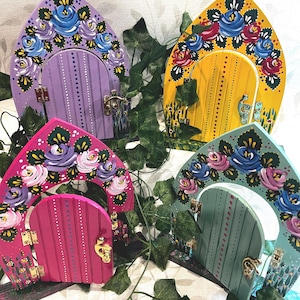 May include: Four arched wooden fairy doors in purple, yellow, pink, and teal, each featuring hand-painted floral motifs in blue, pink, and white along the top arch, vertical slats, gold-toned metal hinges, and a small gold-toned latch handle.