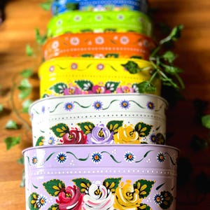 May include: A stack of seven oval metal containers in vibrant colours including pink, blue, green, orange, yellow, white, and lavender. Each container features hand-painted floral motifs with green leaves and small dotted patterns along the rim.