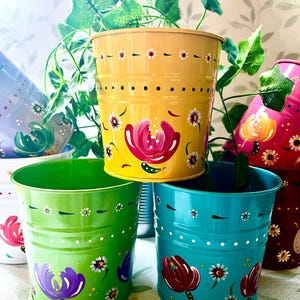May include: A collection of cylindrical metal planters in yellow, green, turquoise, pink, and purple, each featuring hand-painted floral motifs with small white and red blossoms, green leaves, and horizontal dotted lines around the upper rim.