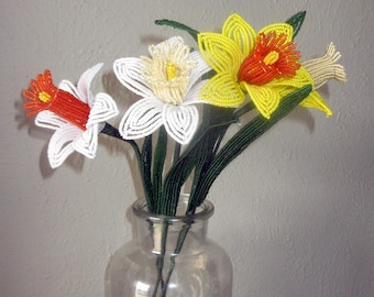 May include: A bouquet of handcrafted beaded flowers in a clear glass vase. The arrangement features three daffodils with white and yellow petals, orange and yellow centers, and long green beaded stems and leaves, all constructed from small glass beads.