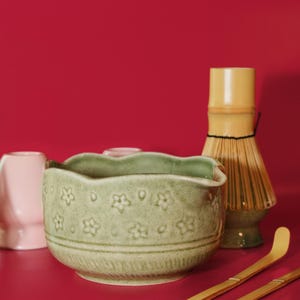 Puede incluir: Un juego de t&eacute; matcha sobre un fondo rojo. El juego incluye un cuenco de cer&aacute;mica verde claro con un dise&ntilde;o floral, un batidor de bamb&uacute;, dos cucharas de bamb&uacute; y una taza de cer&aacute;mica rosa.