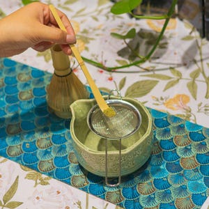 Puede incluir: Preparaci&oacute;n de t&eacute; matcha. Una mano sostiene una cuchara de bamb&uacute; sobre un colador de acero inoxidable, tamizando polvo de matcha verde en un cuenco de cer&aacute;mica verde claro. Tambi&eacute;n se ven un batidor de bamb&uacute; y un camino de mesa estampado.