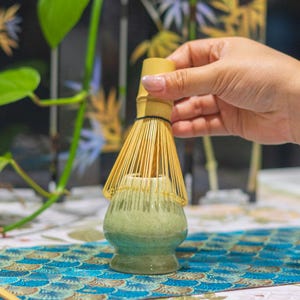 Puede incluir: Un juego de t&eacute; matcha con un batidor de bamb&uacute; y un cuenco de cer&aacute;mica. El cuenco tiene un degradado de verde y azul. El batidor se sostiene sobre el cuenco. El fondo incluye vegetaci&oacute;n y un mantel estampado.