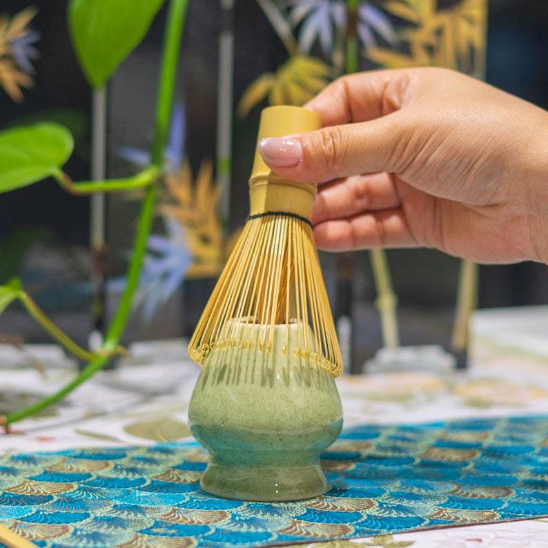 Puede incluir: Un juego de t&eacute; matcha con un batidor de bamb&uacute; y un cuenco de cer&aacute;mica. El cuenco tiene un degradado de verde y azul. El batidor se sostiene sobre el cuenco. El fondo incluye vegetaci&oacute;n y un mantel estampado.