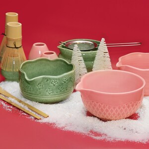 Puede incluir: Colecci&oacute;n de cuencos de cer&aacute;mica y herramientas para la ceremonia del t&eacute; matcha en tonos verdes y rosas, dispuestas sobre una superficie roja con nieve artificial. Incluye un batidor de matcha, cuencos y un colador.