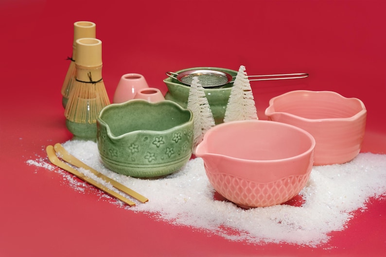 Puede incluir: Colecci&oacute;n de cuencos de cer&aacute;mica y herramientas para la ceremonia del t&eacute; matcha en tonos verdes y rosas, dispuestas sobre una superficie roja con nieve artificial. Incluye un batidor de matcha, cuencos y un colador.