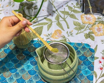 Puede incluir: Un juego de preparación de té matcha con un cuenco de cerámica verde claro, un colador de acero inoxidable y un batidor de bambú. Una cuchara de bambú amarilla contiene polvo de matcha verde. El conjunto está sobre una alfombrilla estampada en azul y dorado, con tela floral en el fondo.