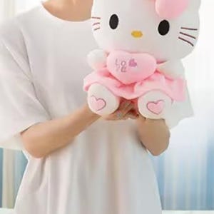 Puede incluir: Un peluche blanco con un personaje felino con ojos negros, bigotes y nariz amarilla. El juguete lleva un lazo rosa, un vestido y sostiene un coraz&oacute;n rosa con la palabra "Love". Los pies del juguete tienen detalles en forma de coraz&oacute;n.