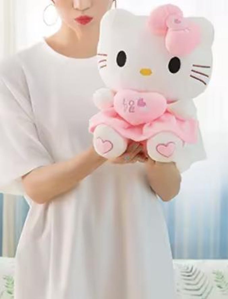 Puede incluir: Un peluche blanco con un personaje felino con ojos negros, bigotes y nariz amarilla. El juguete lleva un lazo rosa, un vestido y sostiene un coraz&oacute;n rosa con la palabra "Love". Los pies del juguete tienen detalles en forma de coraz&oacute;n.