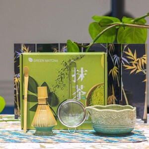 Puede incluir: Un juego de t&eacute; matcha verde con un batidor de bamb&uacute;, una cuchara, un colador y un cuenco de cer&aacute;mica. El juego incluye una caja verde con el texto "Green Matcha" y caracteres japoneses. El fondo presenta una pantalla con tem&aacute;tica de bamb&uacute;.