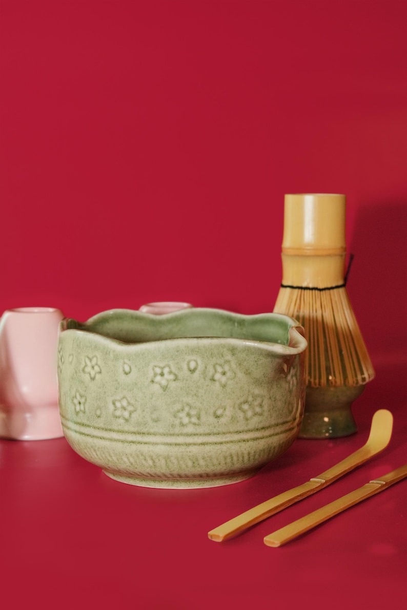 Puede incluir: Un juego de t&eacute; matcha sobre un fondo rojo. El juego incluye un cuenco de cer&aacute;mica verde claro con un dise&ntilde;o floral, un batidor de bamb&uacute;, dos cucharas de bamb&uacute; y una taza de cer&aacute;mica rosa.