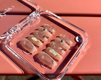Puede incluir: Un juego de diez uñas postizas en un estuche de plástico transparente con la etiqueta XS. Las uñas son de un tono nude translúcido, decoradas con detalles de alambre dorado, pequeñas perlas blancas, dijes de mariposa y piedras de estrás.