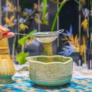 Puede incluir: Una ceremonia de t&eacute; matcha en curso. Una mano sostiene un peque&ntilde;o colador de malla plateada, tamizando polvo de matcha verde en un cuenco de cer&aacute;mica verde claro con dise&ntilde;os florales. Tambi&eacute;n se ven un batidor de bamb&uacute; y un mantel estampado.