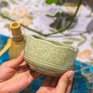 Puede incluir: Un cuenco de t&eacute; de cer&aacute;mica verde claro con un dise&ntilde;o floral y un pico. El cuenco se sostiene con las manos, con un batidor de bamb&uacute; en el fondo. El cuenco tiene una superficie texturizada y un borde festoneado.