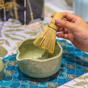May include: A matcha tea set featuring a light green ceramic bowl with floral patterns and a bamboo whisk. The whisk is held above the bowl, ready for use. The set is on a blue and white patterned surface.