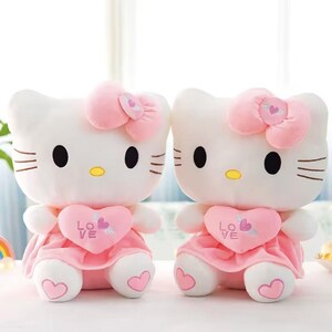 Puede incluir: Dos mu&ntilde;ecas de peluche blancas con forma de gato con ojos negros, narices amarillas y lazos rosas. Cada mu&ntilde;eca lleva un vestido rosa y sostiene un coraz&oacute;n rosa con la palabra "LOVE" y un dise&ntilde;o de flecha. Las mu&ntilde;ecas est&aacute;n sentadas una al lado de la otra.