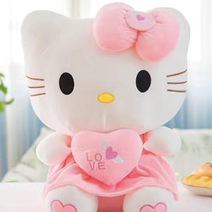 Puede incluir: Un peluche blanco con forma de gato con lazo rosa, vestido y detalle de coraz&oacute;n. El coraz&oacute;n tiene la palabra "LOVE" y un dise&ntilde;o de coraz&oacute;n alado. El gato tiene ojos negros, nariz amarilla y bigotes marrones. Juguete para ni&ntilde;os.