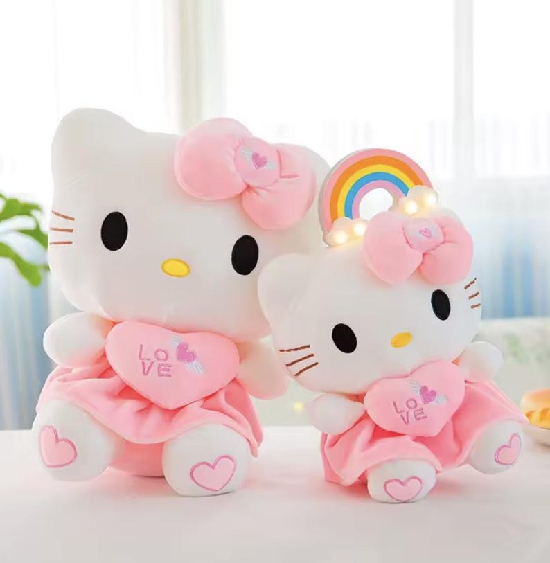 Puede incluir: Dos mu&ntilde;ecos de peluche blancos con forma de gato con lazos y vestidos rosas. Los mu&ntilde;ecos tienen ojos negros, narices amarillas y detalles de coraz&oacute;n rosa. Un mu&ntilde;eco tiene un arco&iacute;ris y luces. Un juguete lindo y tierno.
