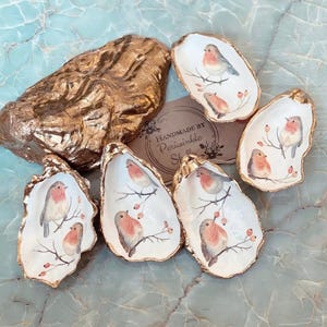 May include: Six gold-painted oyster shells with white interiors, each featuring a hand-painted robin and berries. The text "Handmade by Periwinkle Shells" is visible on a brown tag.