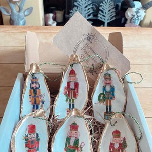 Peut inclure: Six décorations de Noël en forme de casse-noisette, peintes à la main sur des coquilles d'huîtres. Les décorations sont ornées de bordures dorées et présentent différents motifs de casse-noisette. Les décorations sont présentées dans une boîte avec une étiquette indiquant "Handmade Perishable Shells".