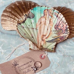 Può includere: Un paio di conchiglie dorate con un'immagine dipinta di una donna in una conchiglia. Le conchiglie sono utilizzate come porta anelli e hanno un'etichetta che dice "Handmade by Periwinkle Shells".