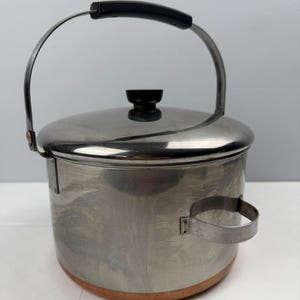 May include: A stainless steel pot with a copper-colored base, featuring a removable lid with a black knob, a tall arched metal handle with a black grip, and a small side handle. The cylindrical body has a polished finish with visible reflections.