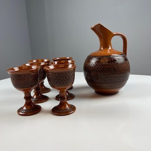 May include: A set of brown ceramic drinkware consisting of a large pitcher with a curved handle and five matching stemmed goblets. Each piece features a dark brown, intricate, embossed geometric lattice pattern around the main body.