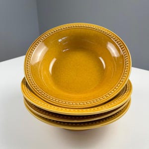 May include: A stack of four round, mustard-yellow ceramic bowls. Each bowl features a smooth, glossy finish and a decorative beaded texture along the outer rim. The bowls are uniform in colour and design, creating a consistent, layered appearance.