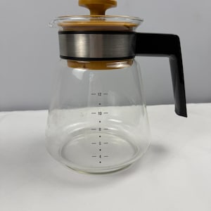 May include: A clear glass coffee carafe with a yellow lid and handle. The carafe has a stainless steel band and black handle. Measurement markings are visible on the side, indicating the volume of liquid in ounces.