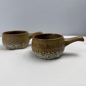 May include: Two brown ceramic bowls with handles, each featuring a textured white speckled base and a band of embossed circular patterns around the upper rim. The bowls have a smooth, glazed finish on the top half and a rough, porous texture below.