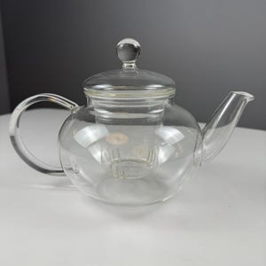 May include: A clear glass teapot featuring a rounded body, a curved handle, a pointed spout, and a removable glass infuser basket inside. The teapot includes a matching glass lid topped with a small, spherical knob handle for easy removal.
