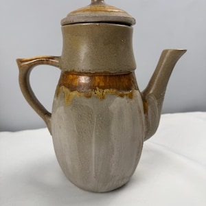 Może przedstawiać: Ceramiczny dzbanek do kawy z pokrywką. Dzbanek jest głównie beżowy z brązowym pasem pośrodku oraz brązowym uchwytem i wylewką. Pokrywka ma mały uchwyt na górze. Dzbanek ma zaokrąglony korpus i teksturowaną powierzchnię.