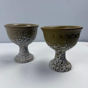 May include: Two ceramic goblets featuring a smooth, olive-toned upper bowl with a subtle etched band near the rim. The lower portion of each goblet and the pedestal base display a thick, white, textured glaze with a rough, cratered appearance.