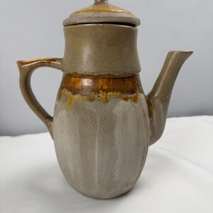 May include: A ceramic teapot with a lid, featuring a two-tone glaze. The upper section and lid are a muted brown, while the lower body is a lighter, textured grey. A distinct band of amber-colored glaze drips between the two color sections.