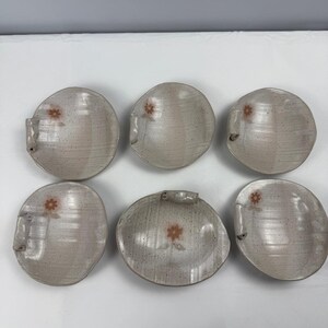 May include: Six speckled, off-white ceramic plates with a small, reddish-brown floral design. Each plate has a unique shape and a small handle. The plates are arranged on a white surface.