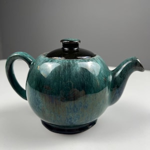 Può includere: Una teiera in ceramica verde acqua e nera con un corpo arrotondato, un manico curvo e un beccuccio. Il coperchio è nero e la teiera ha una finitura lucida. La teiera è progettata per preparare il tè e servire bevande.