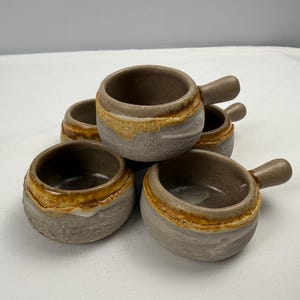 May include: A set of five small ceramic bowls with handles, featuring a textured gray exterior and a smooth, dark brown interior. Each bowl has a distinct, uneven band of golden-brown glaze around the rim, creating a rustic, hand-crafted appearance.