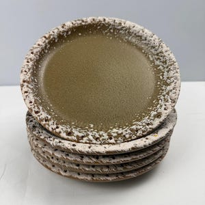 Può includere: Una pila di quattro piatti rotondi in ceramica con un centro verde oliva opaco e liscio e un bordo strutturato e rialzato con macchie bianche. I bordi di ogni piatto presentano un aspetto poroso simile a crateri con un netto contrasto.