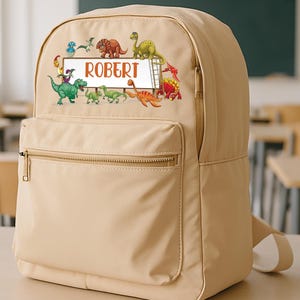 May include: A tan backpack with a front zippered pocket and adjustable straps. The backpack features a colorful dinosaur graphic and the name "ROBERT" on a rectangular label. The backpack is sitting on a light-colored desk in a classroom.