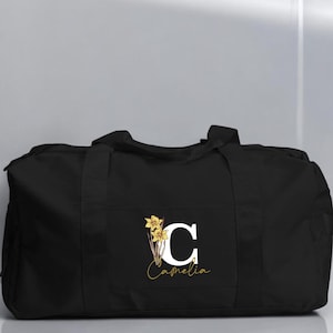 May include: Black duffel bag with a white letter C and the name "Camellia" in gold lettering. The letter C is decorated with a yellow daffodil.