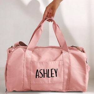 May include: A pink duffel bag with a zipper closure and a front pocket. The pocket has the name "ASHLEY" printed in black.