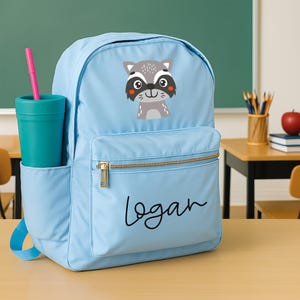 May include: A light blue backpack with a raccoon graphic and the name "logan" in black script. The backpack has a front pocket with a gold zipper and a side pocket holding a teal tumbler with a pink straw.