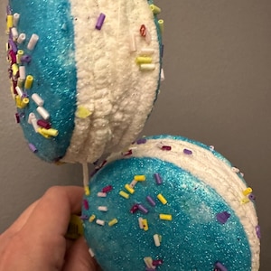 May include: Two decorative ornaments shaped like ice cream sandwiches. Each ornament has a blue glittery half, a white textured half, and colorful sprinkles. The ornaments are held by a hand.