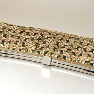 May include: A rectangular clutch with a silver-colored frame and clasp. The clutch features a repeating pattern of beige and dark green floral shapes. The bag is likely made of fabric or a similar material.