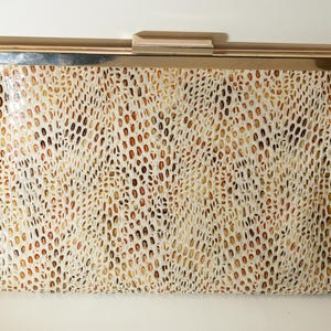 May include: A cream-coloured clutch bag with a textured, reptile-like pattern in shades of brown and tan. The clasp and frame are a shiny, silver-toned metal. This elegant accessory is suitable for formal events.