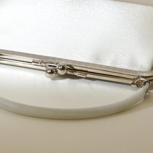 以下が含まれることがあります： シルバーの金属製の留め金と、お揃いのハンドルが付いた白いクラッチバッグ。バッグはテクスチャ加工された表面と丸みを帯びた形状をしています。留め金には、2つの小さなシルバーのボールとヒンジ留めが付いています。