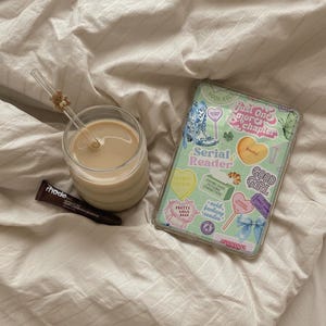 May include: A glass of coffee with a straw, a tube of rhode lip treatment, and a tablet with a decorative case featuring stickers with text like "Serial Reader" and "Good Girl" on a bed.