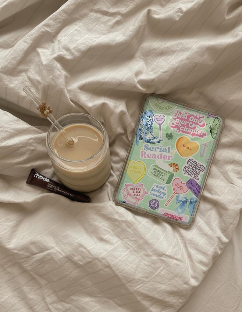 May include: A glass of coffee with a straw, a tube of rhode lip treatment, and a tablet with a decorative case featuring stickers with text like "Serial Reader" and "Good Girl" on a bed.